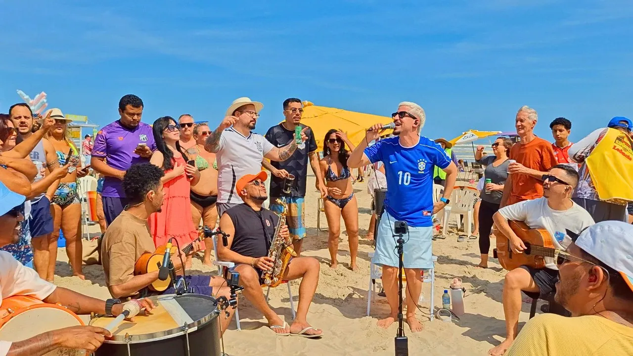 Leleco canta ao microfone em uma roda de pagode na praia, cercado por músicos e público