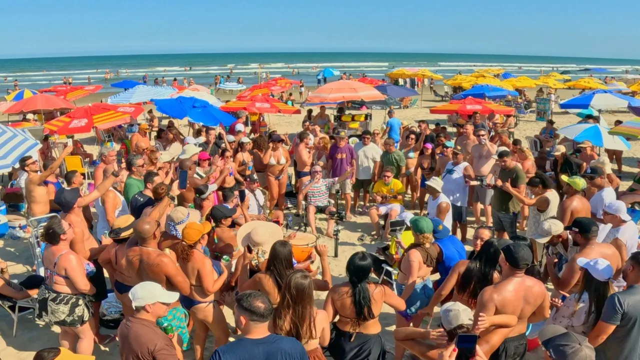 Grande roda de pessoas dançando e cantando ao redor dos músicos, na areia da praia